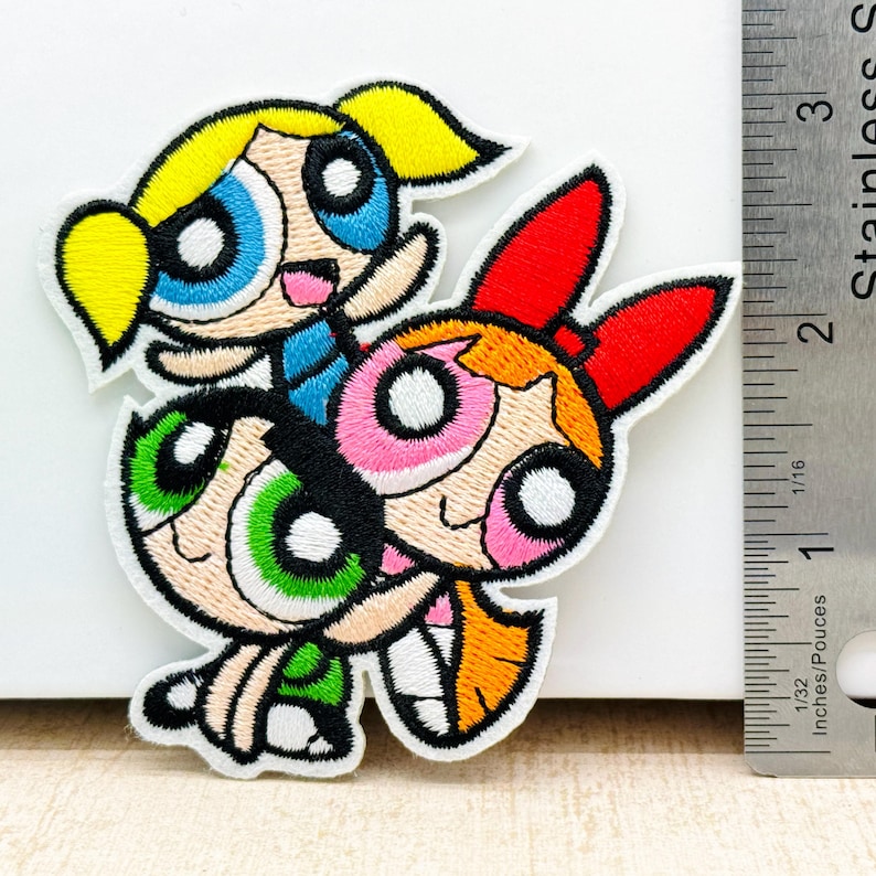 Op de afbeelding: Een geborduurde patch met de Powerpuff Girls, Blossom, Bubbles en Buttercup, in een gestapelde pose. De patch is wit met kleurrijke geborduurde details.