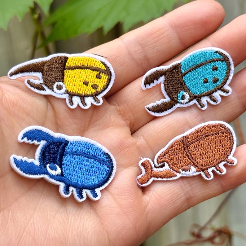 May include: Four embroidered beetle patches in yellow, blue, and brown with white borders. The patches feature detailed stitching and are held in a hand, showcasing the variety of colours and designs.