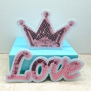 May include: Two decorative patches: a pink sequined crown and the word "Love". The crown is white with pink sequins and a pink outline. The word "Love" is in a cursive font, with pink sequins and a pink outline.