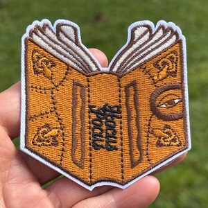 May include: An embroidered patch shaped like an open book with a mustard yellow cover, white pages, and dark brown stitching. The cover features decorative knot patterns, a single eye on the right side, and the text Hocus Pocus in black thread.
