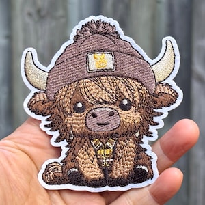 Op de afbeelding: Geborduurde patch met een cartoon Highland-koe met een bruine gebreide muts met een geel vierkant. De koe heeft beige hoorns, een bruine vacht en een kleine gouden kraag. De patch heeft een witte rand.