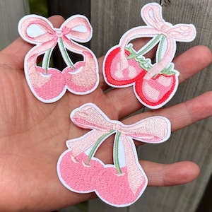 May include: Embroidered patches featuring pink cherries and strawberries with light pink bows and green stems. The patches have a white border and are displayed on a hand.