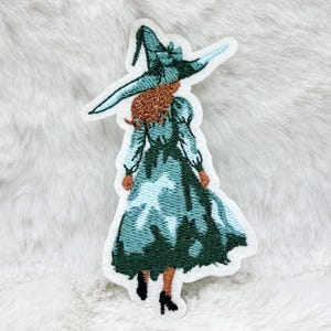 May include: Embroidered patch featuring a witch in a teal dress and hat, viewed from the back. The witch has brown hair and is wearing black heels. The patch has a white border and is on a fluffy white background.