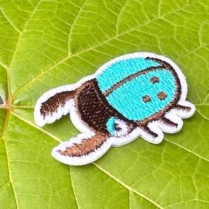 May include: Embroidered patch of a stylized beetle with a turquoise blue top and brown body. The patch has a white border and is set against a green leaf.