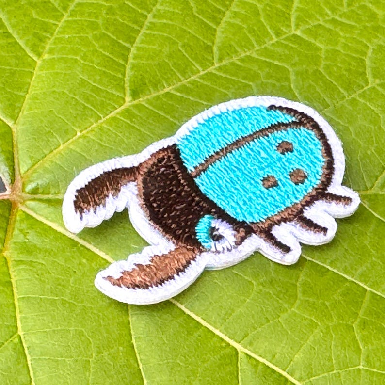 May include: Embroidered patch of a stylized beetle with a turquoise blue top and brown body. The patch has a white border and is set against a green leaf.