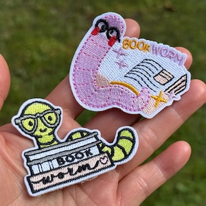 May include: Two embroidered patches featuring bookworm designs. The top patch is a pink worm with black glasses reading a book with the text BOOK WORM. The bottom patch is a lime green worm with black glasses emerging from a stack of books labeled BOOK WORM.