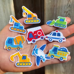 Puede incluir: Colección de parches bordados con varios vehículos. Los parches incluyen una grúa, una hormigonera, un camión de bomberos, un coche de policía, una excavadora, un avión y un camión volquete. Cada parche tiene un borde blanco y colores brillantes.