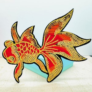 May include: Embroidered patch in the shape of a goldfish. The patch features a red body with yellow and orange accents, and is outlined in black. The design is detailed with sequins and embroidery, creating a textured appearance.