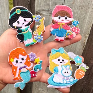 May include: Four embroidered patches featuring cartoon characters: a princess in teal, a girl in a red cape, a mermaid with orange hair, and Alice in a blue dress. Each patch has a unique design with floral and animal accents.