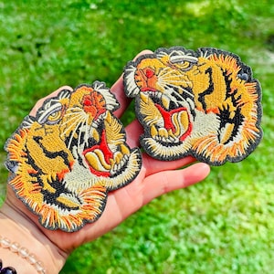 Peut inclure: Deux écussons brodés représentant des têtes de tigre rugissantes. Les écussons présentent un design détaillé avec des fils orange, jaune, noir et blanc. La gueule du tigre est ouverte, révélant des détails rouges et jaunes. Les écussons sont sur fond noir.