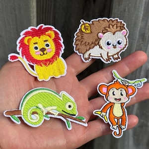May include: Four embroidered fabric patches featuring cartoon animals: a yellow lion with a red mane, a brown hedgehog with a yellow leaf, a green chameleon on a brown branch, and an orange monkey hanging from a green vine with a yellow banana.