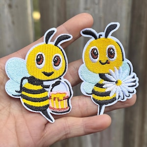 May include: Two embroidered bee patches. One bee holds a honey pot, the other holds a daisy. Both bees have yellow bodies with black stripes, light blue wings, and black antennae. The patches are on a light background.