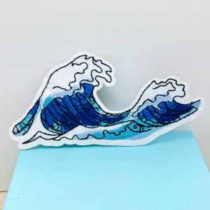 May include: Embroidered patch featuring a stylized ocean wave design. The wave is depicted in shades of blue with white foam, outlined in black. The patch is on a white background and a light blue surface.