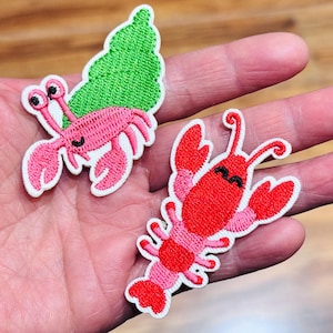 May include: Two embroidered patches featuring cartoon lobsters. One is pink with a green shell, the other is red and pink. Both have white borders and smiling faces. These patches are ideal for adding a nautical touch to clothing or accessories.