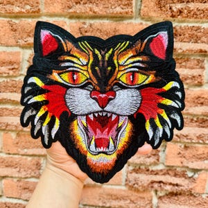 May include: Embroidered patch of a snarling cat's face, with a black background and vibrant details in orange, yellow, red, and white. The cat's eyes are yellow with red pupils, and its mouth is open, revealing white teeth. Suitable for clothing or accessories.