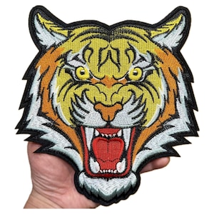 May include: Embroidered tiger head patch with a fierce expression. The patch features a detailed design with yellow, orange, and white colors, and a black border. The tiger's mouth is open, revealing its teeth and a red tongue.