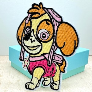 May include: Embroidered patch of a cartoon dog wearing a pink outfit and a pink hat. The dog has brown ears, a yellow body, and a pink nose. The patch is on a light blue background.