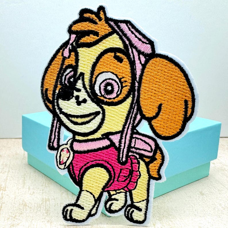 May include: Embroidered patch of a cartoon dog wearing a pink outfit and a pink hat. The dog has brown ears, a yellow body, and a pink nose. The patch is on a light blue background.