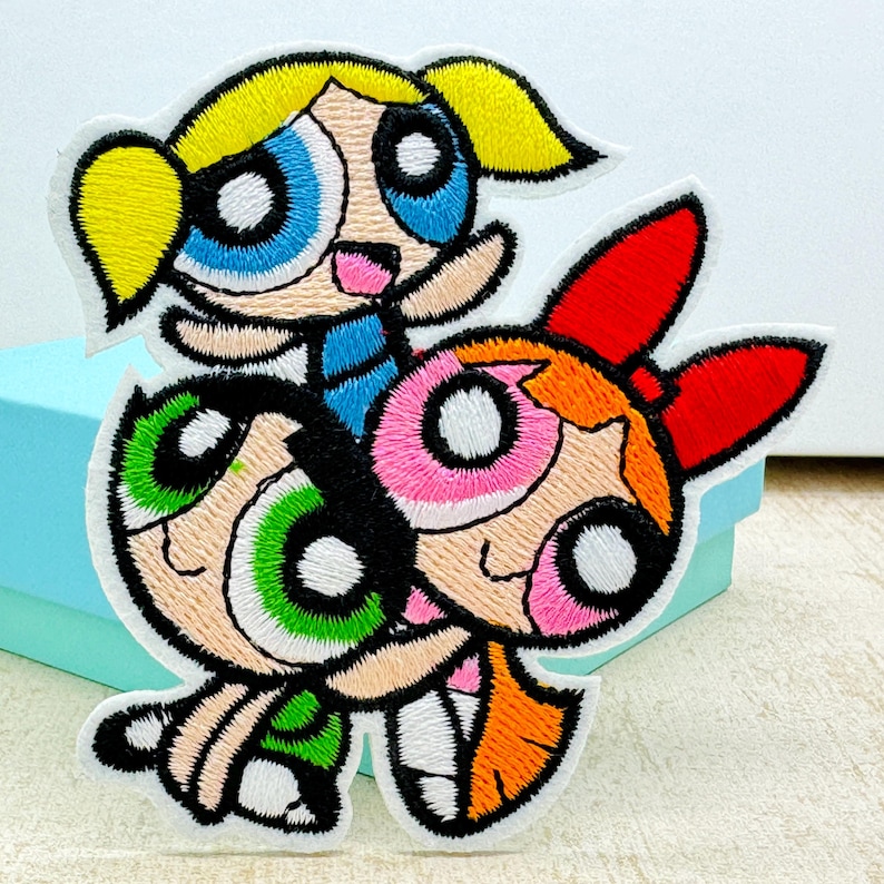 Op de afbeelding: Gestikt patch met de Powerpuff Girls, Blossom, Bubbles en Buttercup. De meisjes zijn afgebeeld in hun kenmerkende poses en kleuren, met Blossom in roze, Bubbles in blauw en Buttercup in groen.