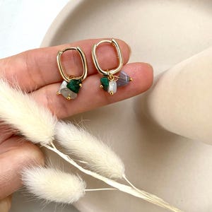 Könnte beinhalten: Goldene Creolen mit einem Anhänger mit drei kleinen Steinen: grün, weiß und lila.