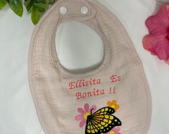 May include: Pink baby bib with a butterfly and flower embroidery. The bib has the text "Ellisita Es Bonita!!" embroidered on it.