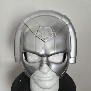 Könnte beinhalten: Ein silberfarbener Helm mit einem markanten Design, der ein zentrales Emblem und ein Visier aufweist. Der Helm wird auf einem weißen Schaumstoffkopf und einem schwarzen Ständer präsentiert, was auf ein Kostüm oder Sammlerstück hindeutet.