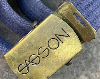May include: A blue fabric belt featuring a rectangular brass-toned metal buckle. The buckle has a brushed finish and displays the word SASSON engraved in black lettering across the front surface. The belt strap is made of a woven textile material.