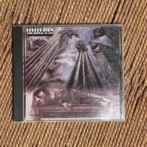 May include: A compact disc case featuring the album cover for Steely Dan's The Royal Scam. The artwork displays a person lying on a bench beneath tall, dark, pointed structures against a cloudy sky. The text Steely Dan and The Royal Scam is visible.