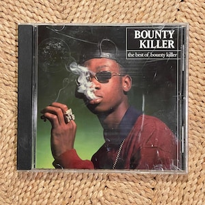 May include: CD case featuring the album "Bounty Killer: The Best of Bounty Killer." The cover art depicts a man in sunglasses smoking, with the album title in a white box. The background has a gradient of green and yellow.