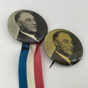 May include: Two circular pinback buttons featuring a black and white portrait of Franklin D. Roosevelt. The larger button on the left includes a vertical ribbon with blue, white, and red stripes. The smaller button on the right has a gold-toned finish.