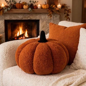 May include: A pumpkin-shaped cushion made of textured, burnt orange sherpa fabric with a black stem, positioned next to a square burnt orange sherpa throw cushion and a white throw cushion on a white textured sofa.