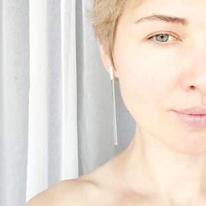 May include: A close-up shot of a person wearing a single, long, minimalist earring. The earring is a thin, straight, clear rod suspended from a small white stud. The background is a soft, white curtain.