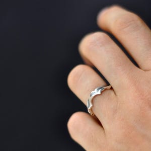 May include: A silver ring with a unique, wave-like design. The ring is worn on a finger, showcasing its intricate, textured surface. The ring's design features a series of curved, pointed elements, creating a distinctive and stylish look.
