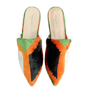 May include: A pair of pointed-toe velvet slippers with a geometric pattern in shades of green, orange, black, and white. The slippers have a white leather insole with the brand name "VELVETICA" printed on them.