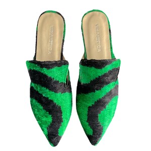 May include: A pair of green and black patterned velvet slippers with pointed toes. The slippers have a simple design and are made of a soft, comfortable material. The brand name "VELVETICA" is printed on the inside of each slipper.