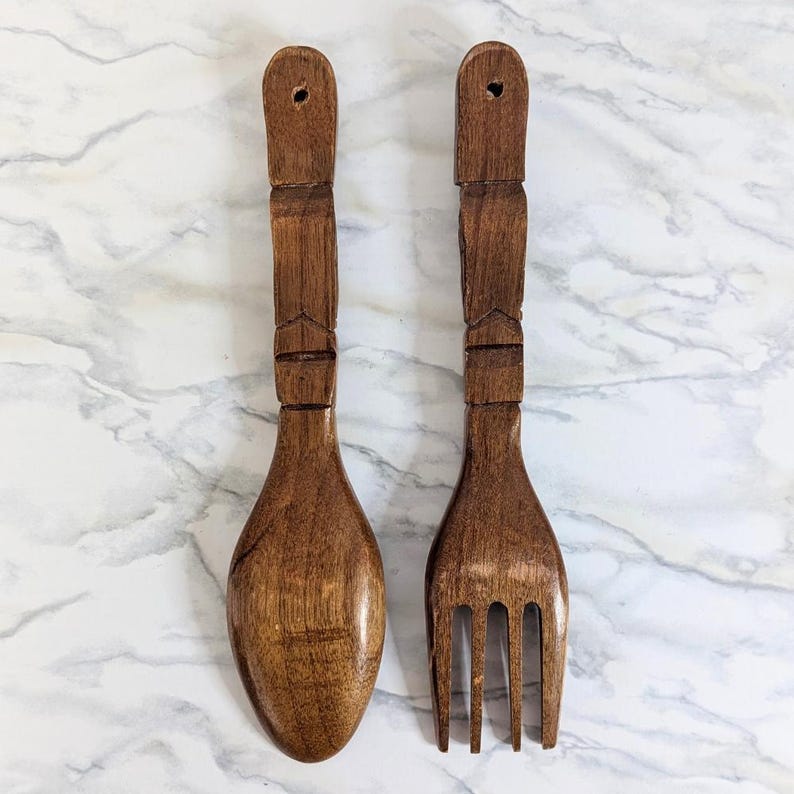 May include: A wooden spoon and fork set. The utensils are made of dark brown wood with carved details on the handles. The spoon has a rounded bowl, and the fork has four tines. Each handle has a hole for hanging.