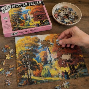 May include: A Guild Picture Puzzle box with a pink border and a 304-piece count, featuring a landscape illustration of a church in autumn. A partially assembled puzzle of the same scene lies below, with loose pieces in a bowl nearby.