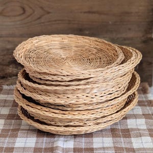 May include: A stack of five round, light brown woven wicker plates featuring a tight, circular coil pattern in the center and a braided edge detail. The plates are constructed from natural reed material with a smooth, uniform finish throughout.