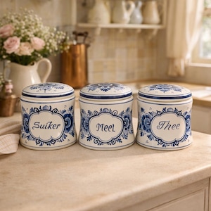 May include: Three ceramic canisters with lids, featuring a blue and white floral design. The canisters are labeled "Suiker", "Meel", and "Thee". The lids and bases are trimmed with blue bands. The canisters are displayed on a light-colored countertop.