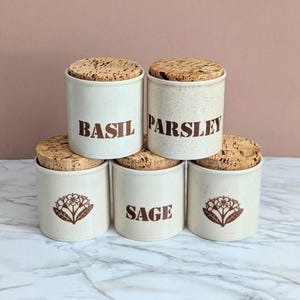 May include: A set of five cylindrical ceramic spice jars with speckled off-white glaze and natural cork lids. Two jars are labeled BASIL and PARSLEY in brown text, one is labeled SAGE, and two feature a brown floral graphic illustration.