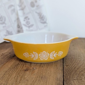May include: A vintage, yellow Pyrex bowl with white floral designs. The bowl has a rounded shape with two handles and a white interior. The floral pattern is repeated around the exterior of the bowl. The bowl is sitting on a wooden surface.