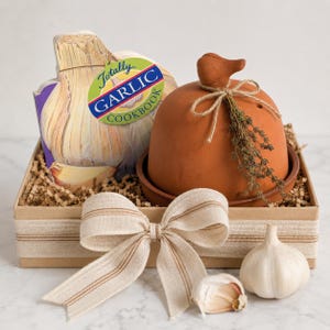 May include: A gift basket featuring a garlic cookbook, a terracotta garlic baker, and fresh garlic bulbs. The basket is decorated with a cream-colored bow and contains shredded paper for presentation. The cookbook has the text "Totally Garlic Cookbook".