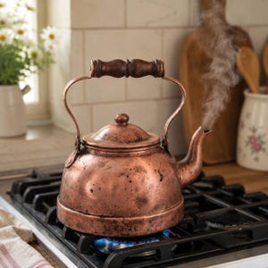 May include: A copper tea kettle with a wooden handle is steaming on a gas stove. The kettle is a warm, reddish-brown color and has a classic design. Steam is escaping from the spout, indicating the water is boiling. The stove is black, and the background includes kitchen items.