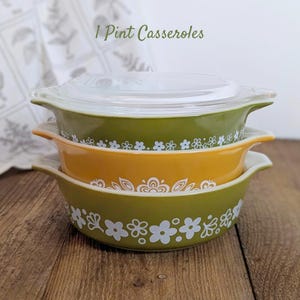 May include: Three vintage 1-pint casseroles stacked, featuring a clear lid on top. The casseroles are in shades of green and yellow, adorned with white floral patterns. The text "1 Pint Casseroles" is visible at the top. The casseroles are on a wooden surface.