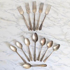 May include: An assortment of vintage silverware, including forks and spoons, arranged on a marble-patterned surface. The silverware has a tarnished, antique appearance, suggesting age and use. The arrangement is symmetrical, with forks at the top and spoons below.