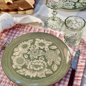 May include: A green ceramic plate with a floral design, a clear glass, and a wine glass are set on a pink and white checkered tablecloth. A knife and fork are also visible. The plate features a cream-colored floral pattern.