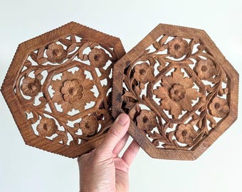 May include: Two octagonal wooden trivets featuring intricate hand-carved floral patterns. Each piece displays a central flower surrounded by smaller blossoms and leafy vines within a geometric frame, showcasing a natural medium brown wood grain.