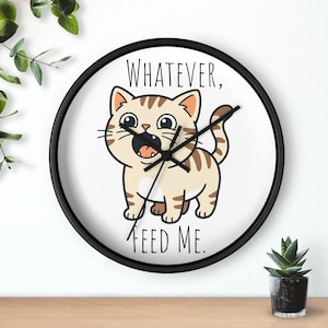May include: A round wall clock with a black frame. The clock face features a cartoon cat with the text "WHATEVER, FEED ME." The cat is beige with brown stripes and an open mouth. Black clock hands indicate the time.