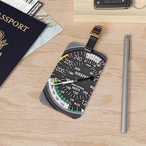 May include: A rectangular luggage tag featuring a black and white aviation airspeed indicator graphic with white numbers and text including AIRSPEED, TRUE SPEED, KNOTS, and M.P.H. The tag has a black leather strap with a gold-toned metal buckle.