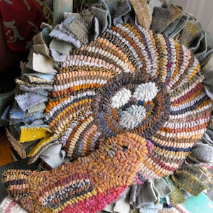 Puede incluir: Una alfombra circular hecha a mano con un diseño de pájaro. La alfombra presenta un patrón en espiral en tonos marrones, beige y naranja, con una aplicación de pájaro en colores similares. Los bordes están adornados con retazos de tela.